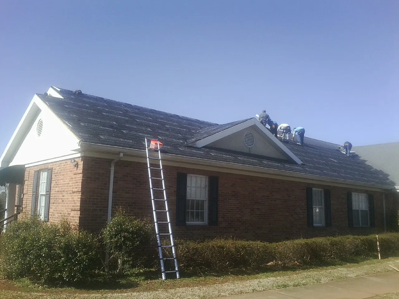 Crew tearing shingles off a brick ranch roof with a ladder set — Emergency Roof Repair work by our crew in Ballico