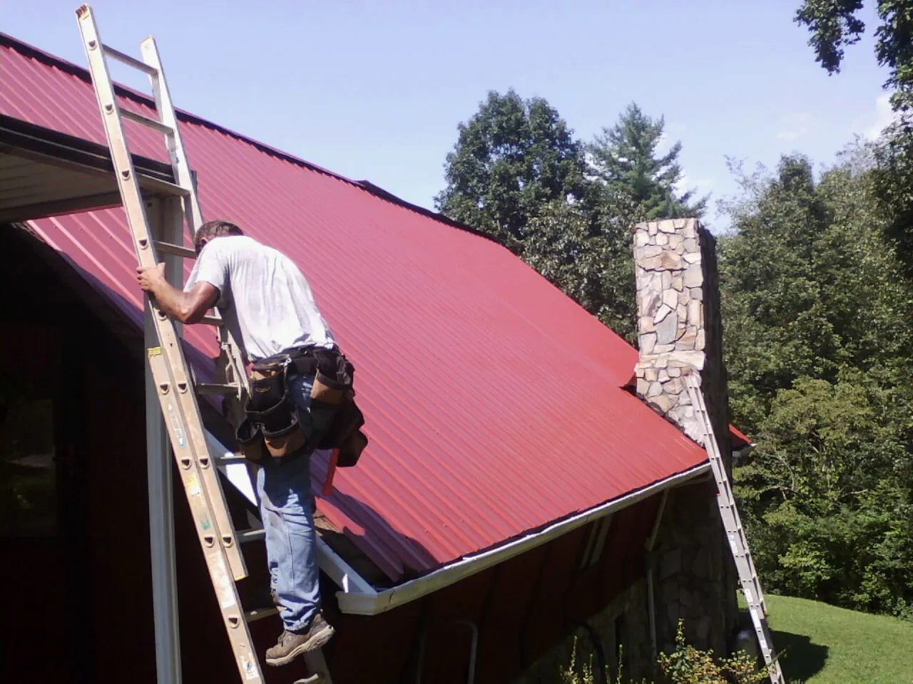 Roofer on a ladder beside a red metal roof and stone chimney — Soffit & Fascia Repair by Roofing Brothers in Ballico