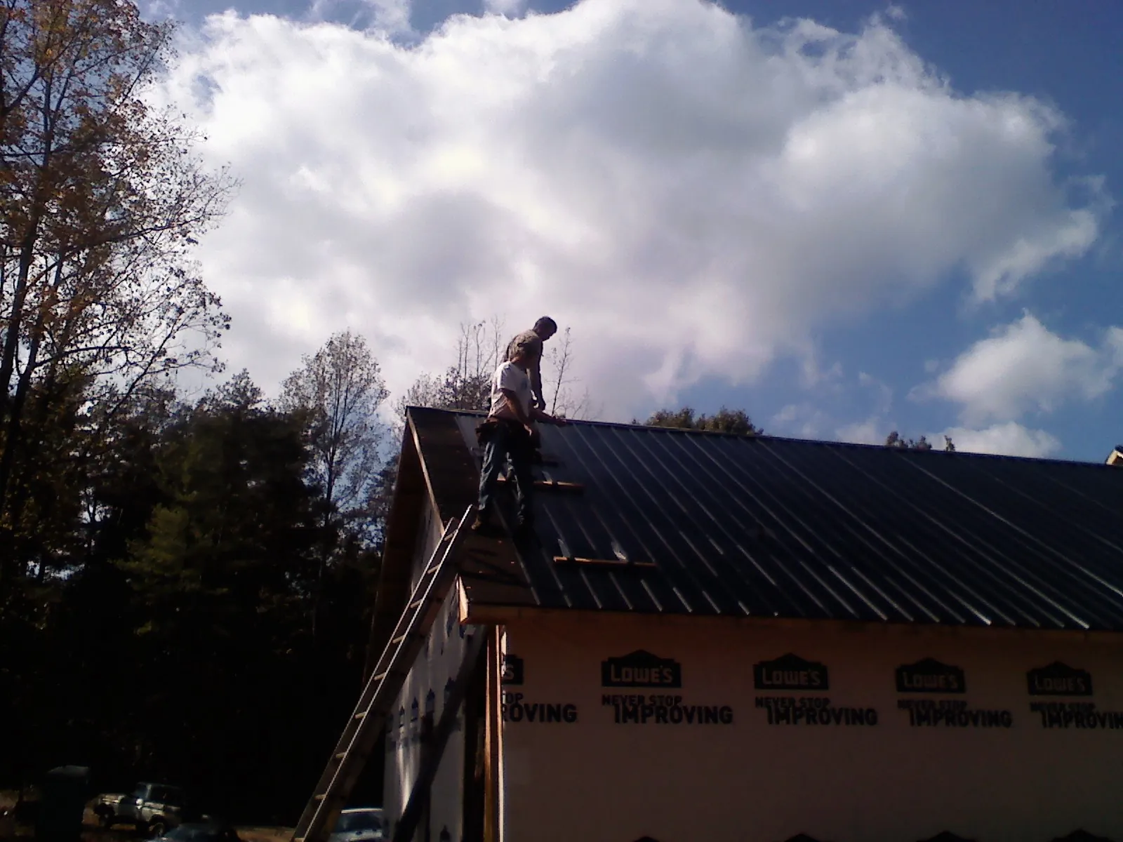 Roofer fastening dark metal panels high against a cloudy sky, from a Roof Inspection job in Ballico